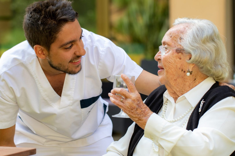 Close up portrait of senior woman with young male nurse outdoors. Man with arm on woman’s shoulder. Old woman with home assistant in garden.