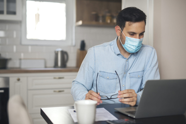 Young man sitting in front of laptop working remotely and wearing mask due to virus pandemic. Man wearing mask due to virus pandemic.