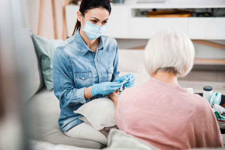 Physician in medical face mask holding syringe while senior lady keeping arm on cushion stock photo Female doctor sitting on couch with old woman